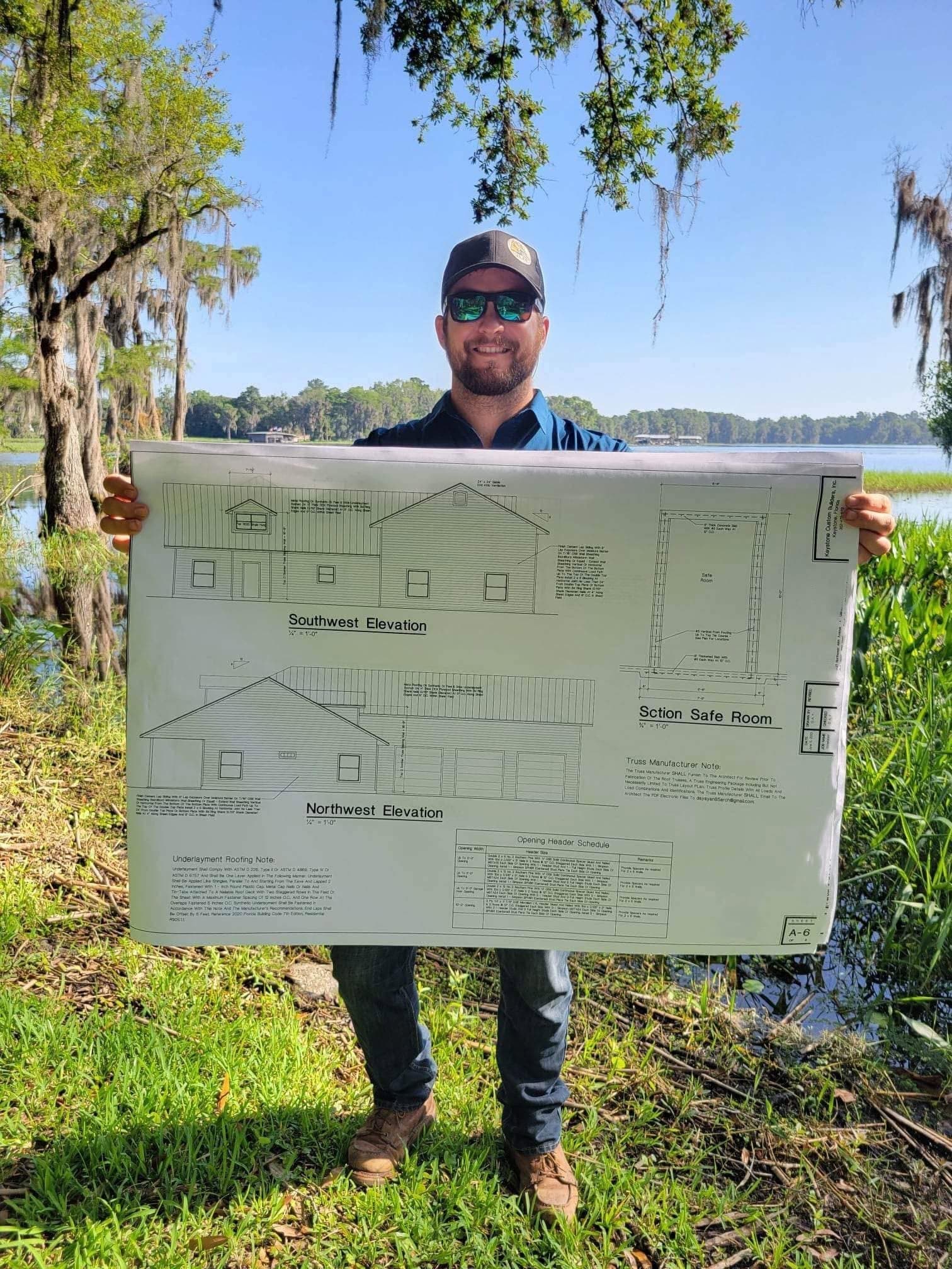 Robert Johnson of Keystone Custom Builders holding custom home elevation plans on a wooded lakefront lot
